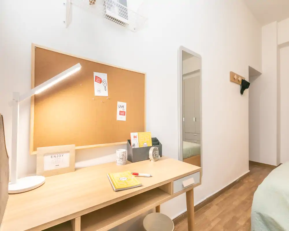 Rincón de dormitorio luminoso y ordenado con escritorio de madera, tablero de corcho y vista parcial de la cama. El espacio está bien iluminado y acondicionado para una zona de estudio o junto a la cama.