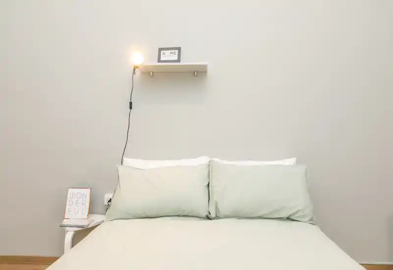 Dormitorio luminoso y minimalista con una cama doble bien hecha, ropa de cama en tonos pastel y una pequeña estantería con lámpara que crea un ambiente tranquilo y acogedor.