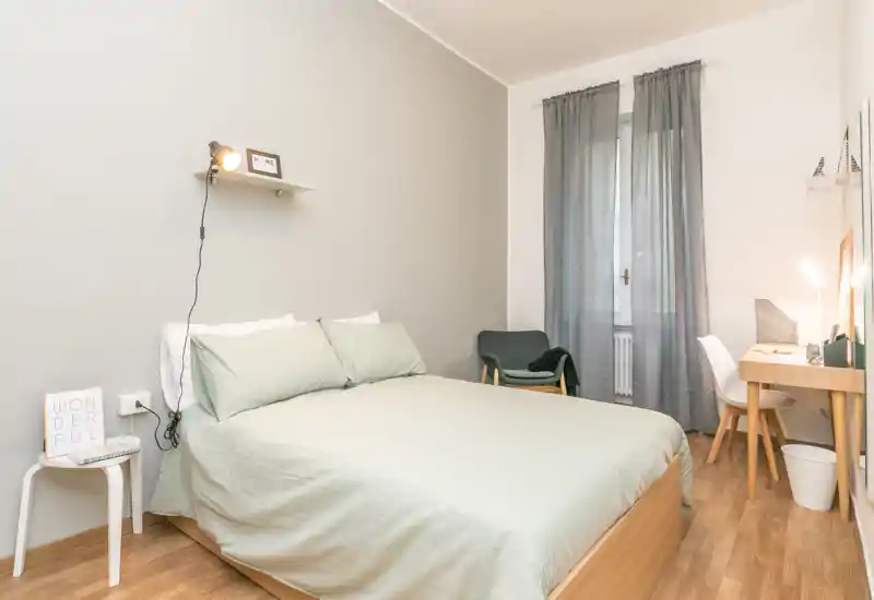 Dormitorio luminoso y ordenado con cama doble vestida en verde claro, una butaca junto a la ventana y un escritorio compacto — ideal para alquileres de corta estancia.