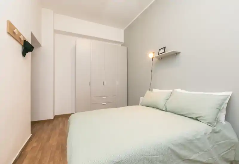 Dormitorio ordenado y bien iluminado con cama de matrimonio vestida con ropa de cama verde claro, armario sencillo y lámpara de lectura en la pared — ideal para un anuncio de alquiler que destaque comodidad y estilo limpio.