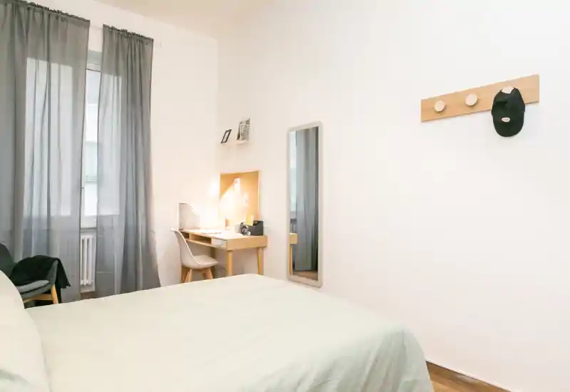 Dormitorio luminoso y minimalista con la cama bien hecha, un escritorio de madera pequeño y un espejo de cuerpo entero; la luz natural filtrada por las cortinas genera una atmósfera tranquila.