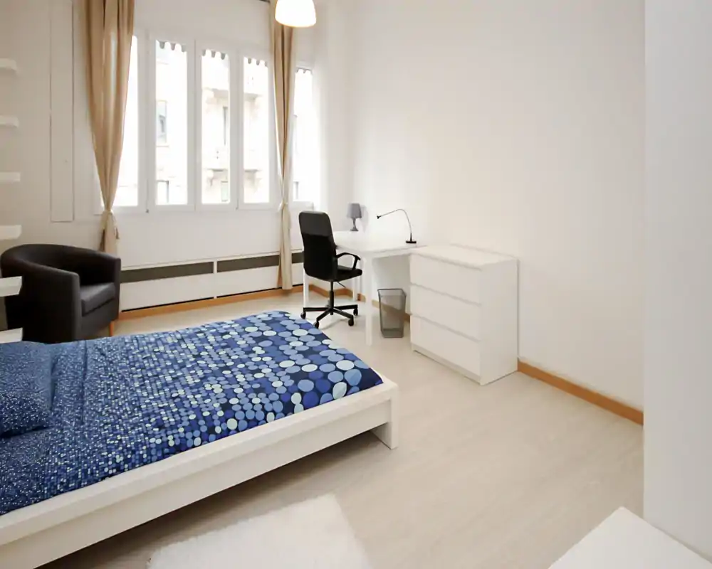 Bright, minimal bedroom with a white platform bed dressed in blue patterned bedding, a comfortable armchair, and a small workspace by large windows letting in abundant natural light.