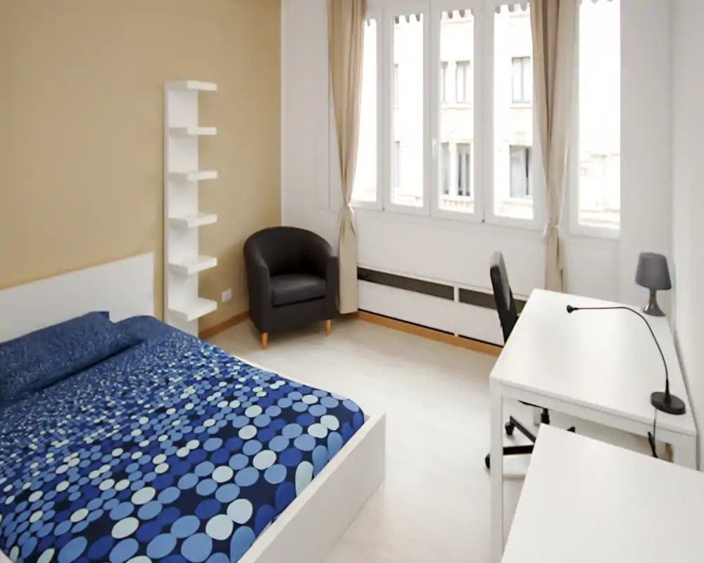 Bright, modern bedroom with a double bed dressed in a blue patterned cover, a comfortable armchair, and a white desk by large windows letting in abundant natural light.