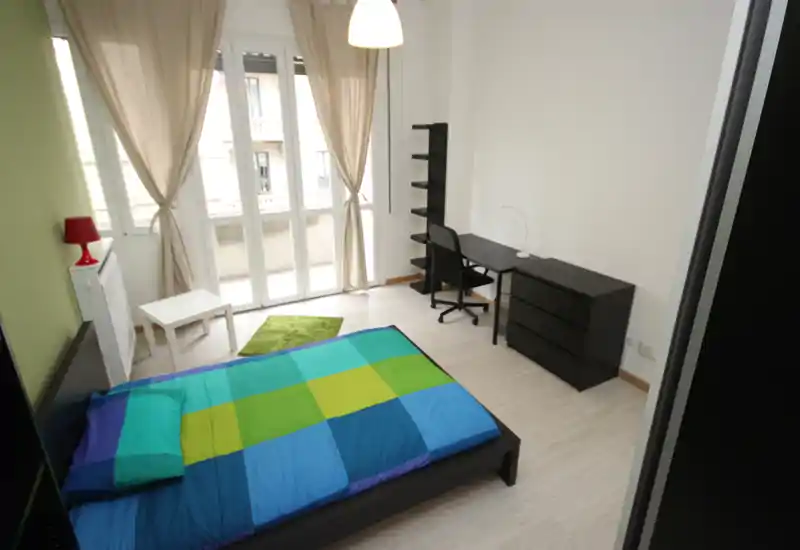 Bright, modern bedroom with a double bed, desk and shelving by large French-style windows letting in abundant natural light.
