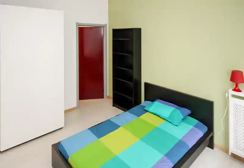 Bright, modern bedroom with a double bed featuring colorful bedding, a black headboard, open shelving and a bedside lamp. Clean, minimal styling and pleasant natural light make it ideal for rental photos.