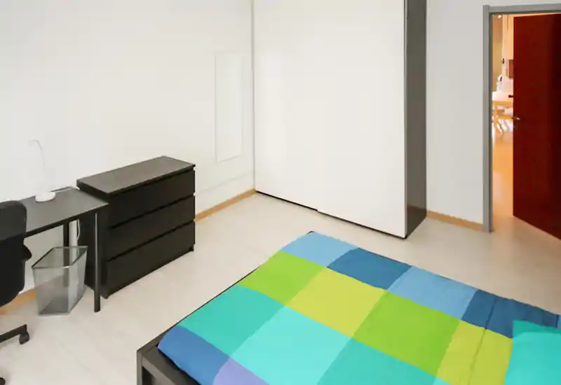 Bright, minimalist bedroom with a colorful double bed, black desk and chest of drawers; simple, clean lines and good natural light.
