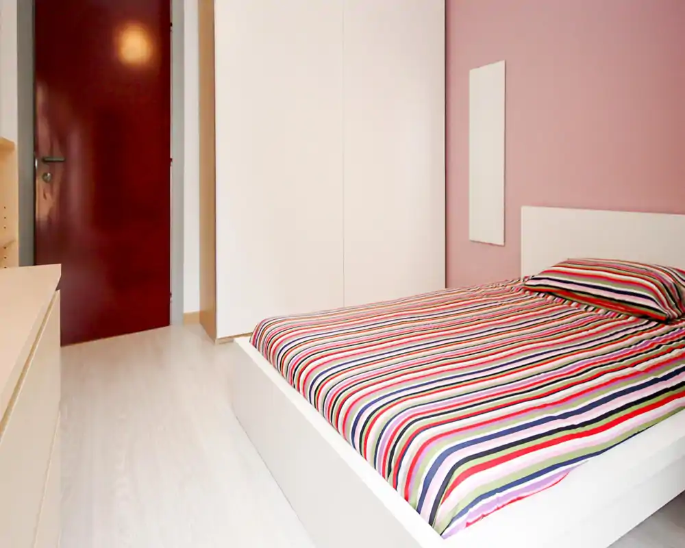 Dormitorio moderno y luminoso con cama plataforma blanca y ropa de cama a rayas de colores, gran armario y suelo de madera clara—limpio y luminoso.