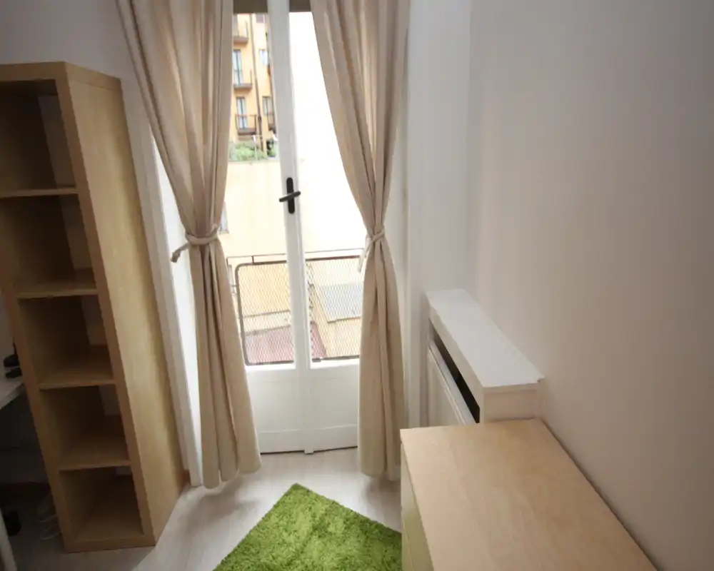 Espacio de estar compacto con una puerta de vidrio a un pequeño balcón, cortinas neutras y una alfombra verde. Estantería y un pequeño mueble de madera clara ofrecen almacenamiento en una habitación luminosa y sencilla.