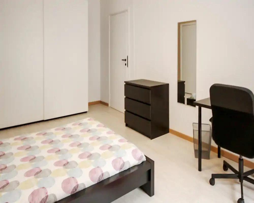 Camera da letto luminosa e minimal con letto basso in primo piano, copripiumino a fantasia, comò nero, scrivania e sedia — uno spazio pulito e confortevole ideale per studenti o professionisti.