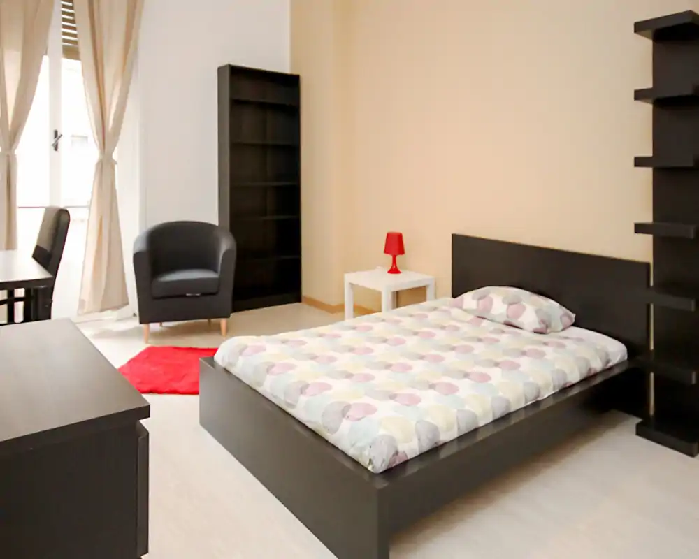 Camera da letto moderna e luminosa con letto matrimoniale, comodino con lampada, poltrona e scaffali; toni neutri e pavimento chiaro donano un'atmosfera ariosa e minimalista.