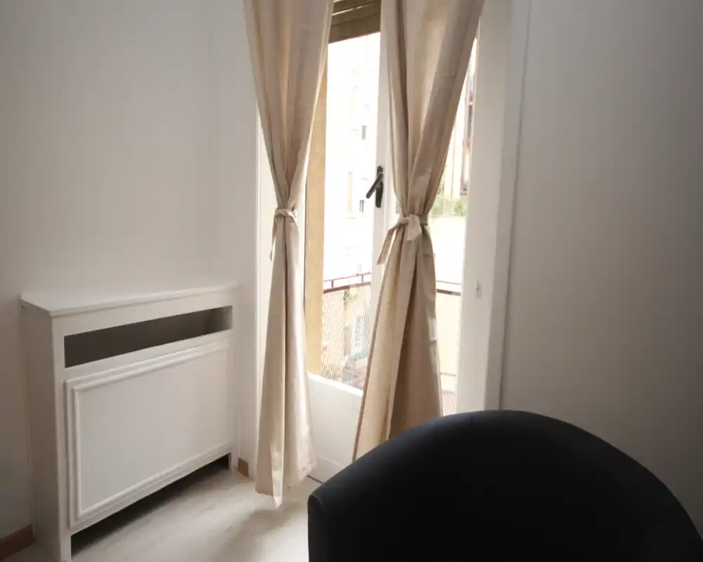 Piccola zona giorno luminosa con finestra coperta da tende e una poltrona scura imbottita; toni neutri e luce naturale creano un'atmosfera tranquilla.