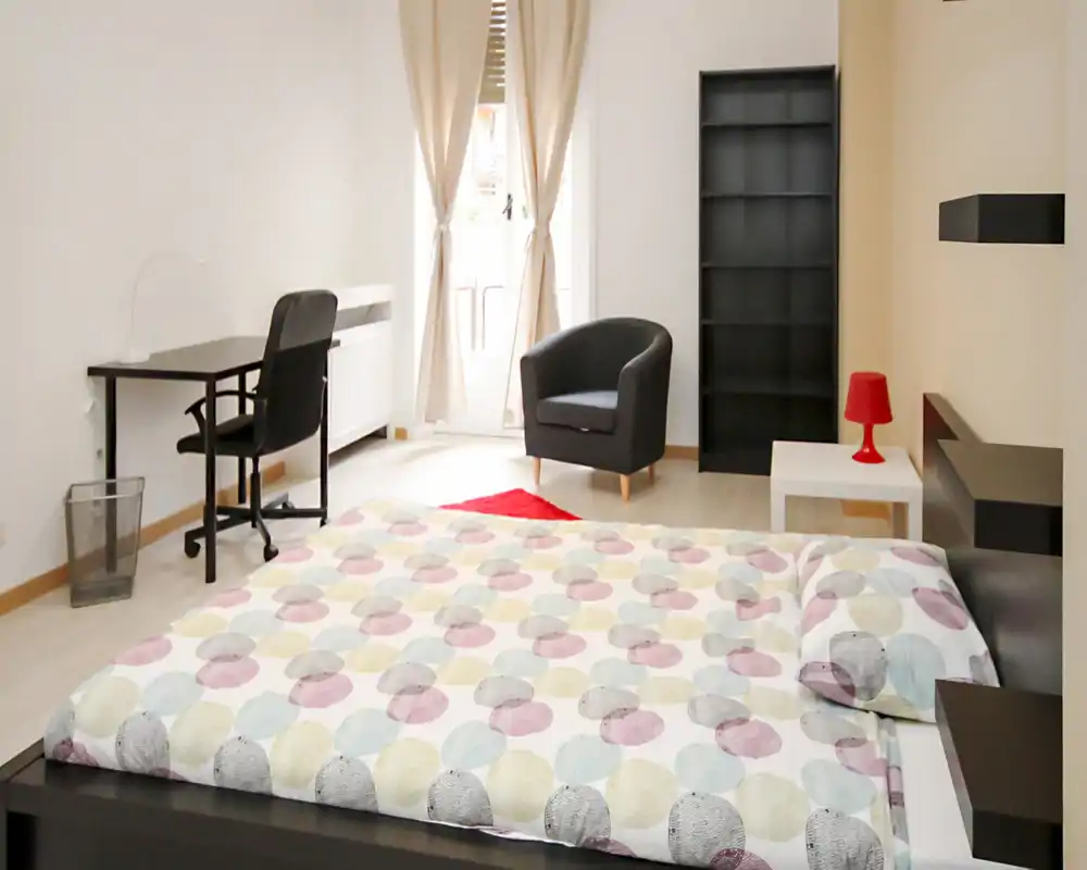 Camera da letto luminosa e moderna con letto matrimoniale in primo piano, una scrivania con sedia, una poltrona comoda e scaffalatura; tessuti morbidi e luce naturale creano un'atmosfera accogliente.