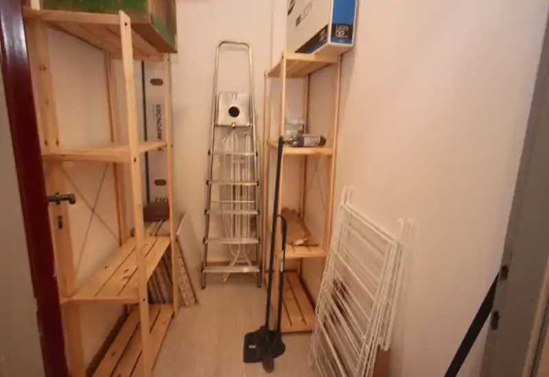 Small storage/utility room with wooden shelving, a folding ladder, cleaning tools and a drying rack. Functional space for boxes and household items.