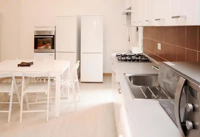 Bright modern kitchen with a built-in oven, two refrigerators, gas hob, stainless sink and a dining table with chairs; clean minimalist styling suitable for rental photos.