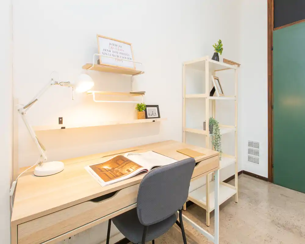A bright, minimal study corner with a wooden desk, adjustable desk lamp, open book and a comfortable chair beside a shelving unit — ideal for focused work or reading.