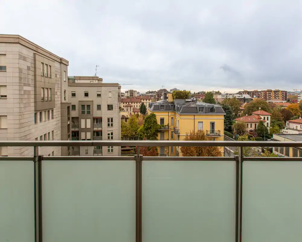 Balcony with frosted glass railing offering an urban view of neighboring buildings and treetops under a cloudy sky.