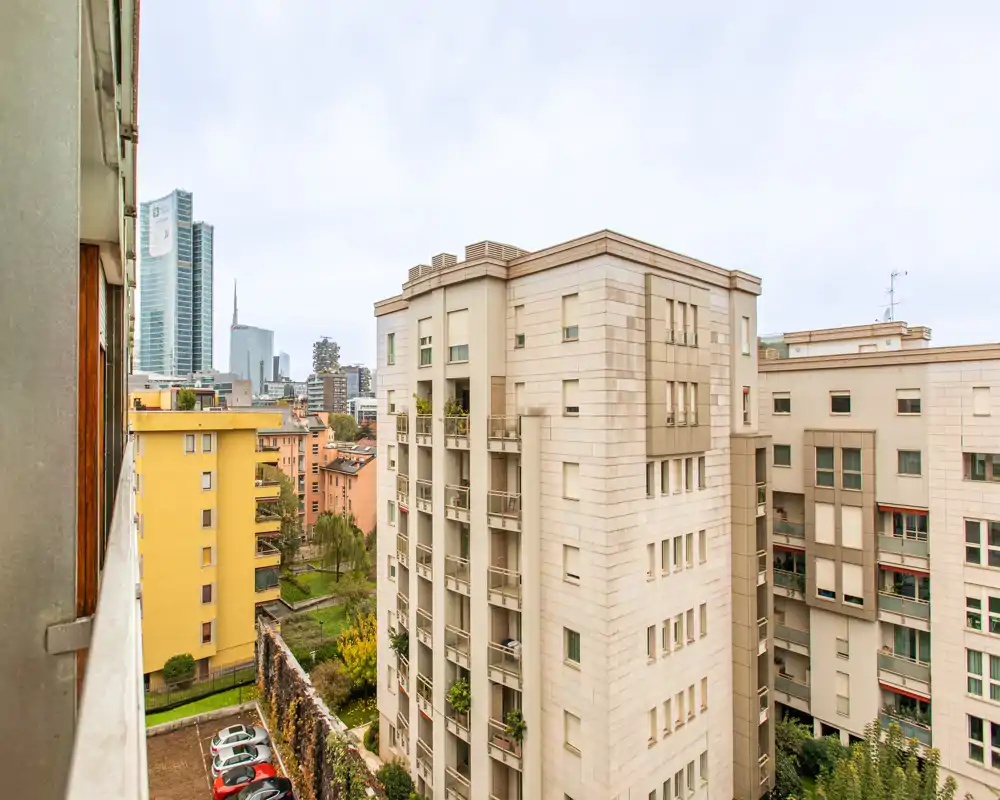 Urban balcony/terrace view showing neighboring apartment buildings, a courtyard with parked cars and a distant skyline — ideal to illustrate location and surroundings.