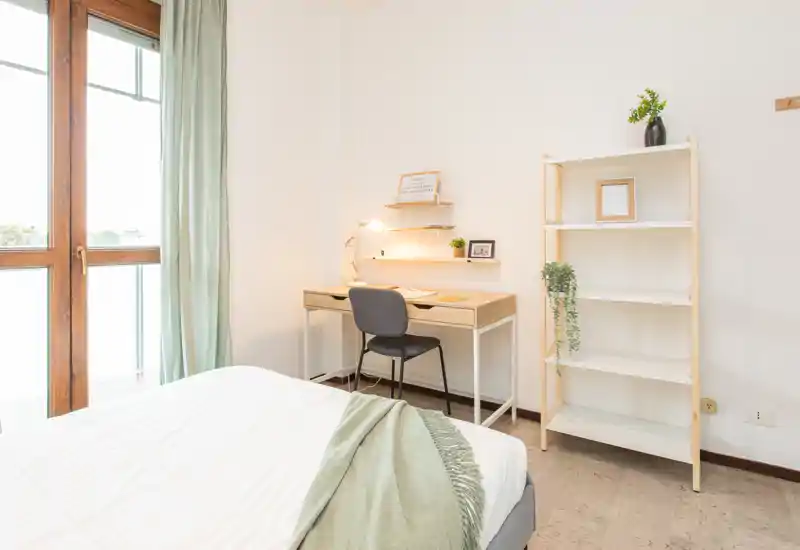 Bright, minimalist bedroom with a comfortable bed, bedside textiles and a small desk by the window offering natural light.