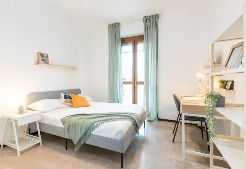 Bright, modern bedroom with a double bed dressed in white linens, soft green throw and yellow accent pillow; natural light from a tall window and a small desk make it ideal for work and relaxation.