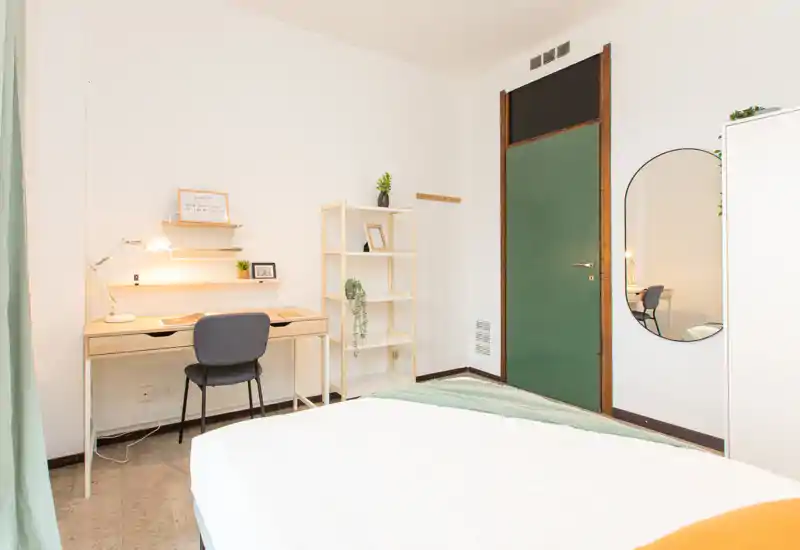 Bright, neatly styled bedroom with a visible double bed in the foreground, a tidy desk and shelving, and soft natural light creating a welcoming atmosphere.