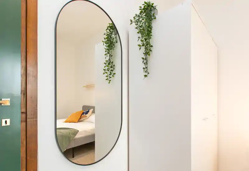 A stylish bedroom view focused through an oval wall mirror; you can see a neatly made bed with cushions and a throw, soft lighting and simple modern decor.