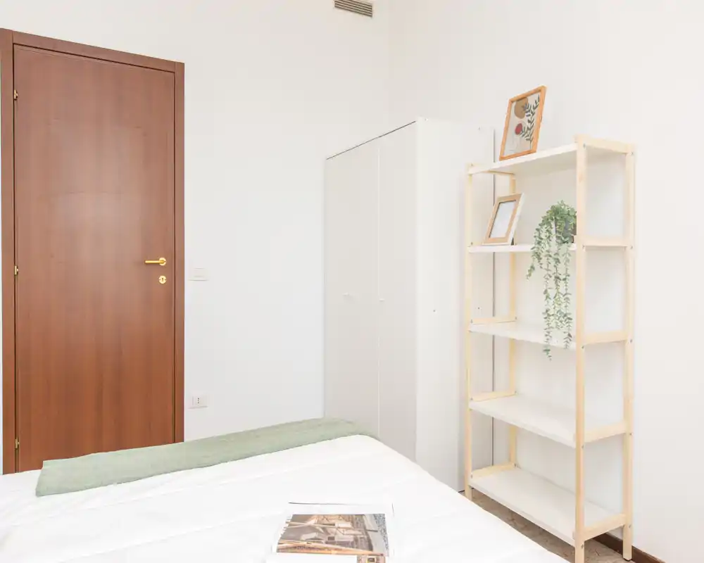 Bright, minimal bedroom with a neatly made bed, a white wardrobe and a wooden shelving unit; clean neutral palette and natural light make it welcoming for guests.