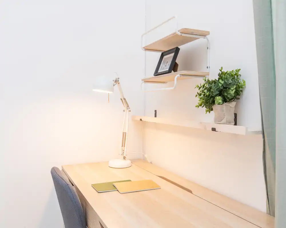 Bright, minimalist corner workspace with a wooden desk, adjustable desk lamp, shelving, a framed photo and a small plant — ideal for focused work or study.