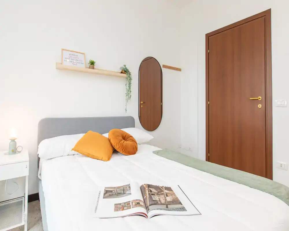 Bright, neatly staged bedroom with a queen bed, decorative mustard cushions, a bedside table lamp and wall mirror — ideal for relaxing stays.