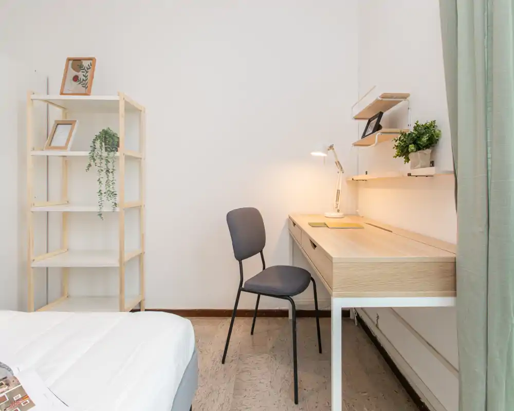 Clean, minimal bedroom corner with a visible bed, a wooden desk with lamp and chair, and shelving with plants—bright and simply styled.