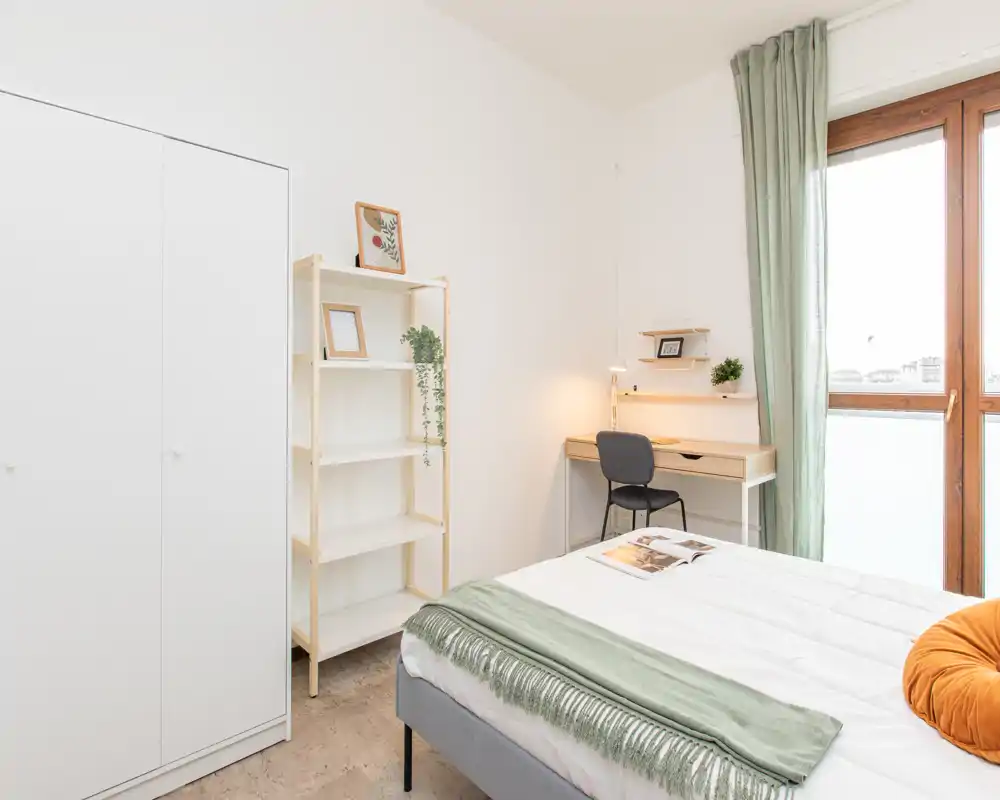Bright, minimalist bedroom with a comfortable double bed, soft green accents, a small desk by the window and a wardrobe — ideal for a calm, well-lit stay.