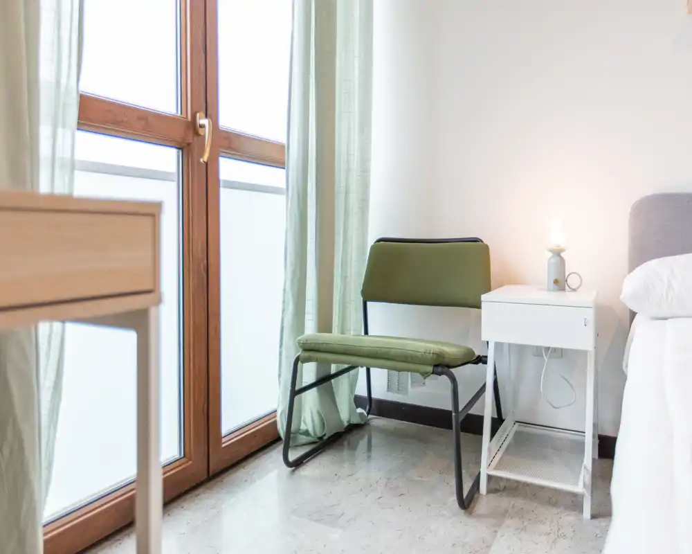 Bright, minimal bedroom corner with a bedside table, modern chair and part of a white bed by a large window letting in soft natural light.