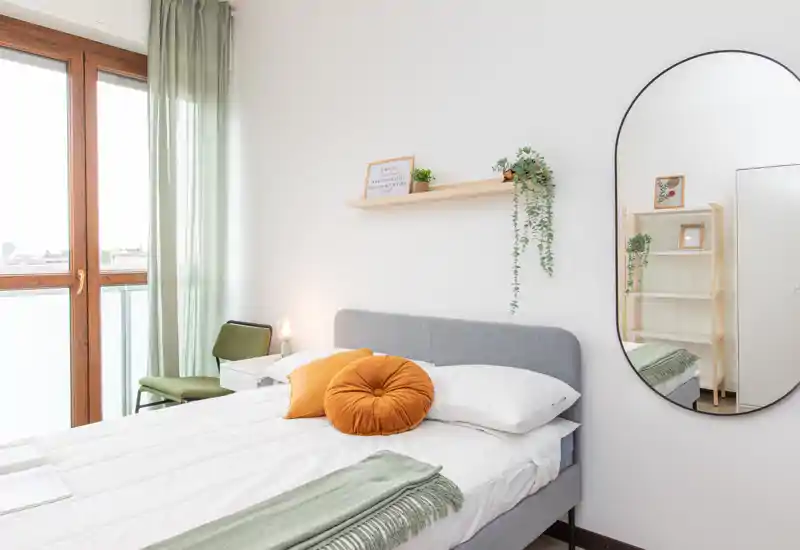 Bright, modern bedroom with a neatly made bed, decorative cushions and natural light from a large window. A round wall mirror and subtle plants create a calm, welcoming atmosphere ideal for rental listings.
