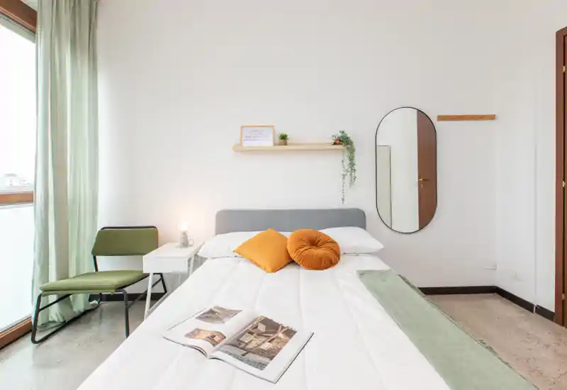 Bright, minimalist bedroom with a neatly made double bed, decorative cushions, a bedside table with lamp, and a large window that fills the room with daylight.