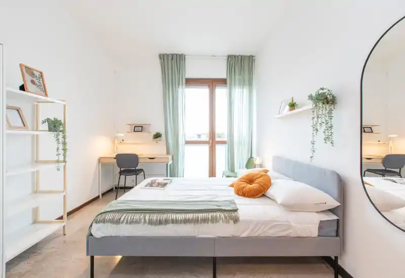 Bright, modern bedroom with a neatly made double bed, soft green accents and natural light from a large window. A small desk and shelving provide workspace and storage, while decorative plants and a round cushion add cozy touches.