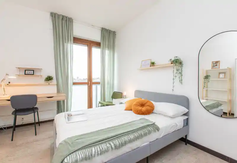 Bright, styled bedroom with a double bed, green accents and a work desk by the window. Large window with sheer curtains provides natural light and a full-length mirror adds depth.