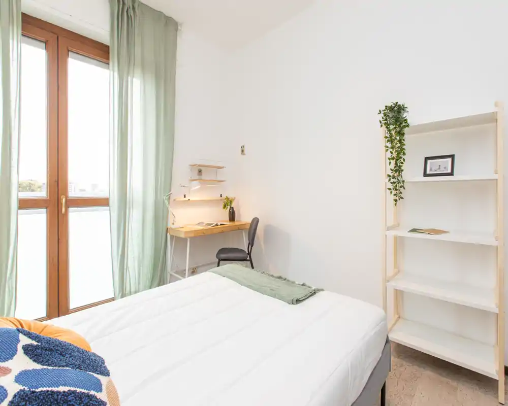Bright, minimal bedroom with a large window, white bedding and a small workspace. The room feels fresh and well lit, with simple shelving and subtle green accents.