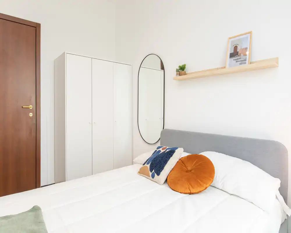 Bright, minimalist bedroom with a neatly made double bed, decorative cushions and a wardrobe. The space features a tall oval mirror and a simple floating shelf for added style.