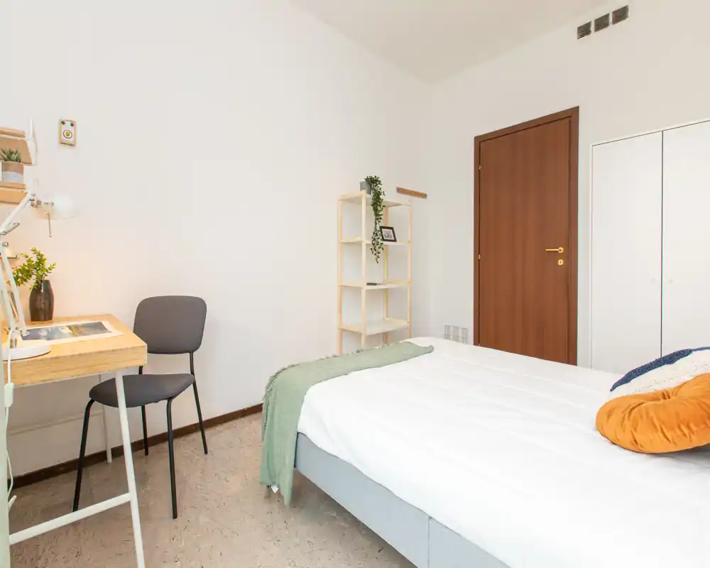 Bright, minimal bedroom with a neatly made double bed, a small desk and chair, and a shelving unit — ideal for a tidy single or couple.