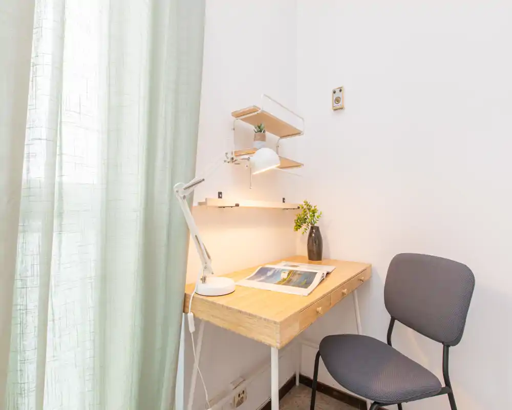 Small, bright workspace corner with a wooden desk, adjustable desk lamp, floating shelves and a simple chair next to a window with light curtains.