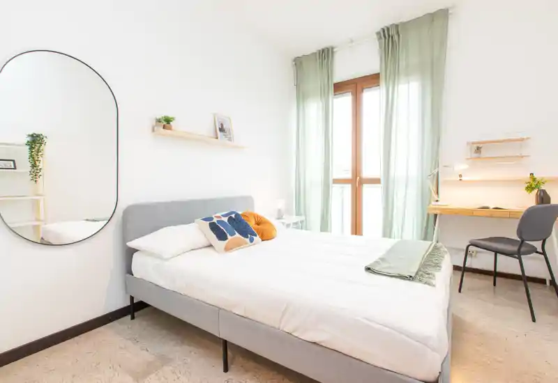 Dormitorio moderno y luminoso con cama doble, cojines decorativos, espejo de pared y un pequeño escritorio junto a la ventana soleada.