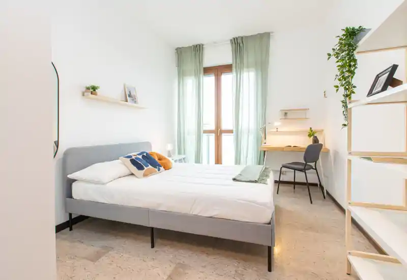 Dormitorio luminoso y moderno con cama totalmente visible, ropa de cama suave, cojines decorativos y un pequeño escritorio junto a la ventana — ideal para estancias cómodas.