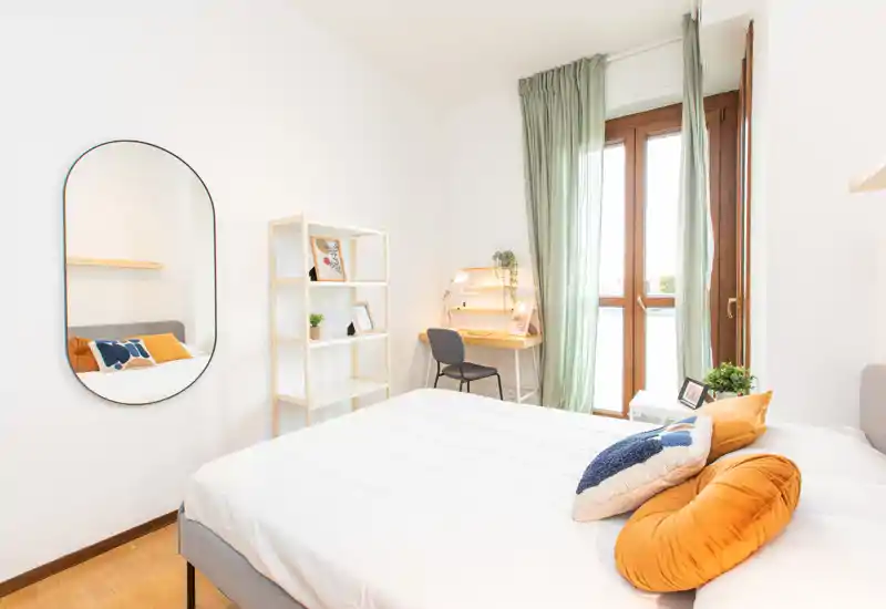 Dormitorio luminoso y bien decorado con cama doble, cojines decorativos y un pequeño escritorio junto a la ventana que aporta mucha luz natural.