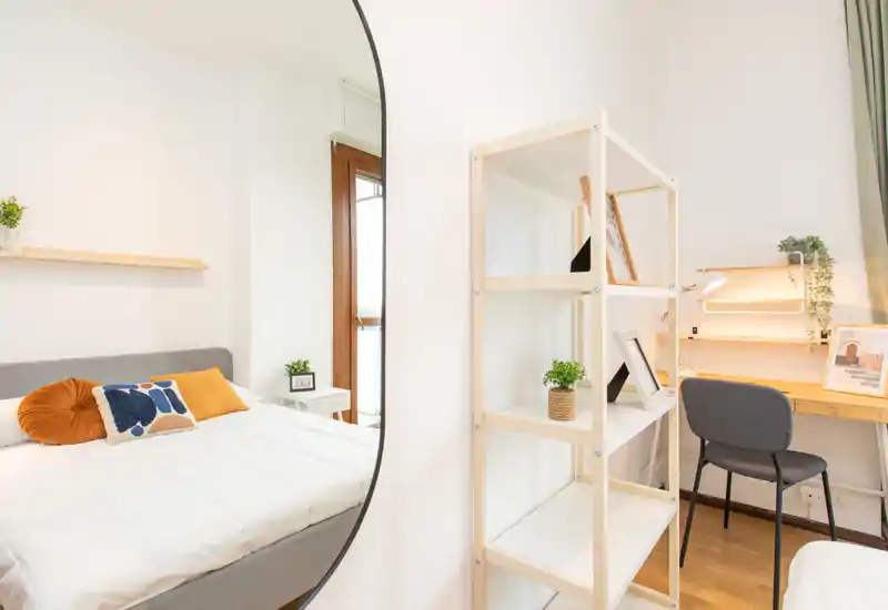 Dormitorio moderno y luminoso con cama doble cómoda, cojines decorativos y rincón de estudio con estantería y escritorio; un gran espejo aporta profundidad y luz natural.