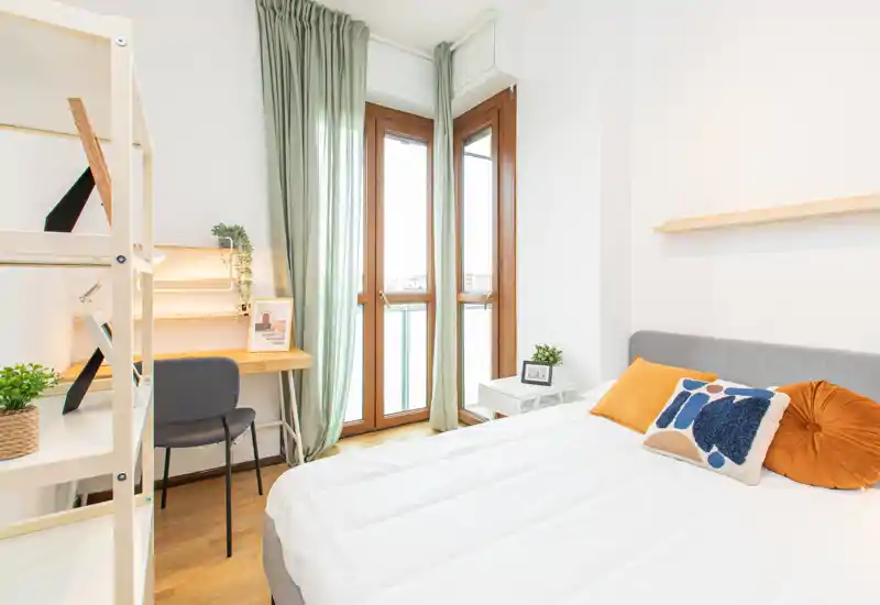 Dormitorio moderno y luminoso con cama doble cómoda, cojines decorativos y un pequeño puesto de trabajo junto a la ventana. Grandes ventanas con marcos de madera y cortinas verdes translúcidas que aportan mucha luz natural; suelos de madera clara y una decoración minimalista crean un ambiente sereno.
