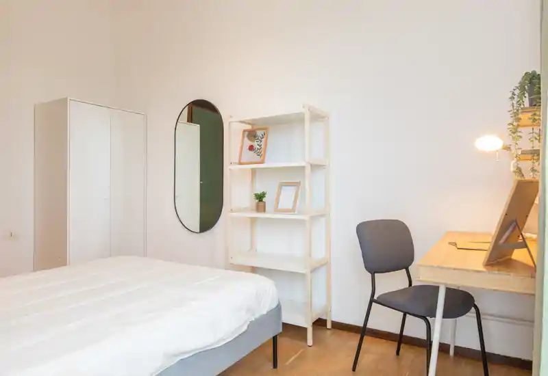 Dormitorio minimalista y luminoso con cama bien hecha, armario, estantería y un pequeño escritorio con luz cálida — sencillo, moderno y acogedor.
