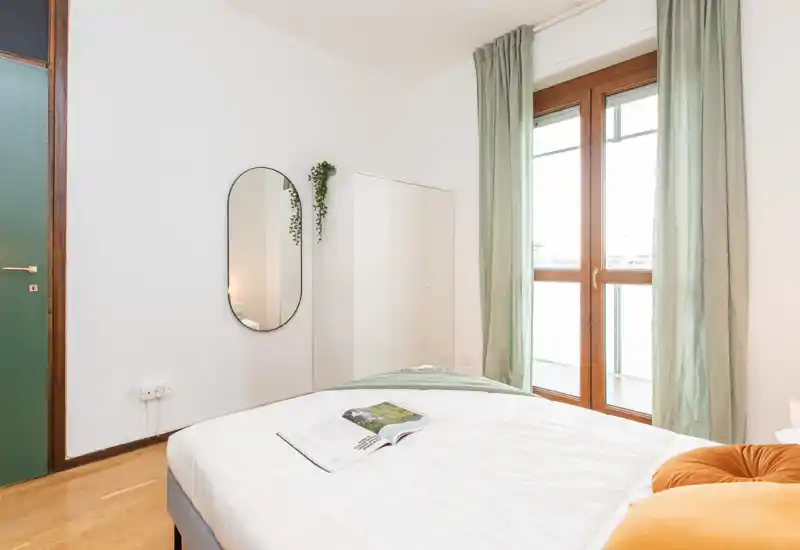 Camera da letto luminosa e minimal con un comodo letto matrimoniale, tessuti soffici e una grande finestra con accesso al balcone. Toni neutri e arredi semplici creano un’atmosfera calma e ariosa, perfetta per annunci.