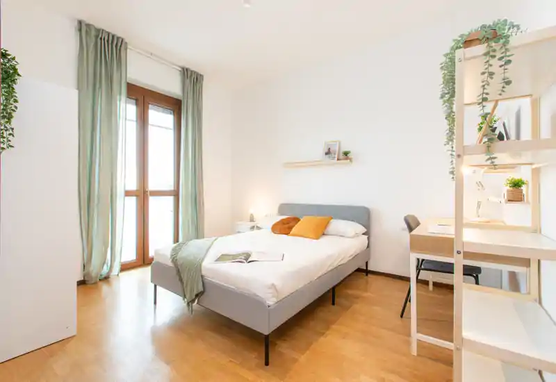 Camera da letto luminosa e ben arredata con letto matrimoniale, comodino e zona studio; ampia finestra che garantisce molta luce naturale e arredi nei toni neutri.