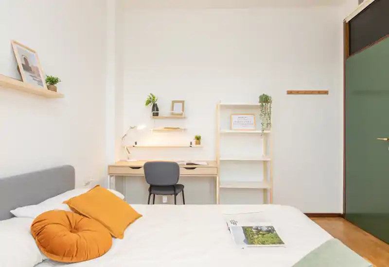 Camera da letto moderna e luminosa con un letto comodo in primo piano, cuscini caldi e una scrivania ordinata con mensole — uno spazio ben arredato, accogliente e adatto sia al riposo che al lavoro.