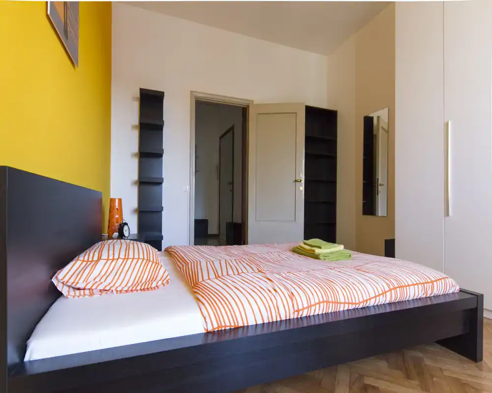 Camera da letto luminosa e moderna con letto basso in legno scuro, biancheria a righe arancioni e parete accentuata gialla; scaffali e armadio offrono spazio pratico.