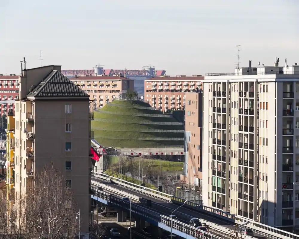 Vista urbana luminosa che mostra palazzi residenziali vicini, una strada sopraelevata e un tumulo verde terrazzato in lontananza — perfetta per valorizzare la posizione e il contesto cittadino.